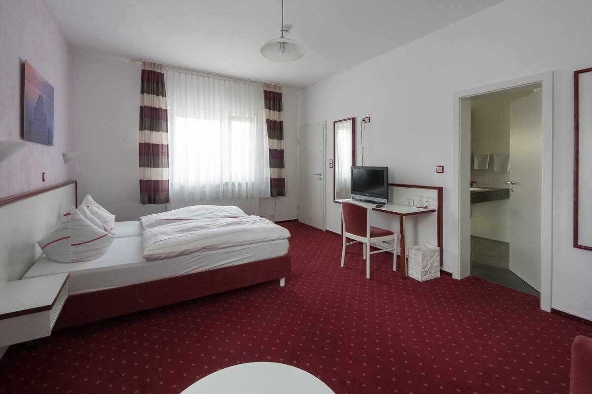 A hotel room with a bed, a desk with a computer, a mirror, and a bathroom visible through an open door.