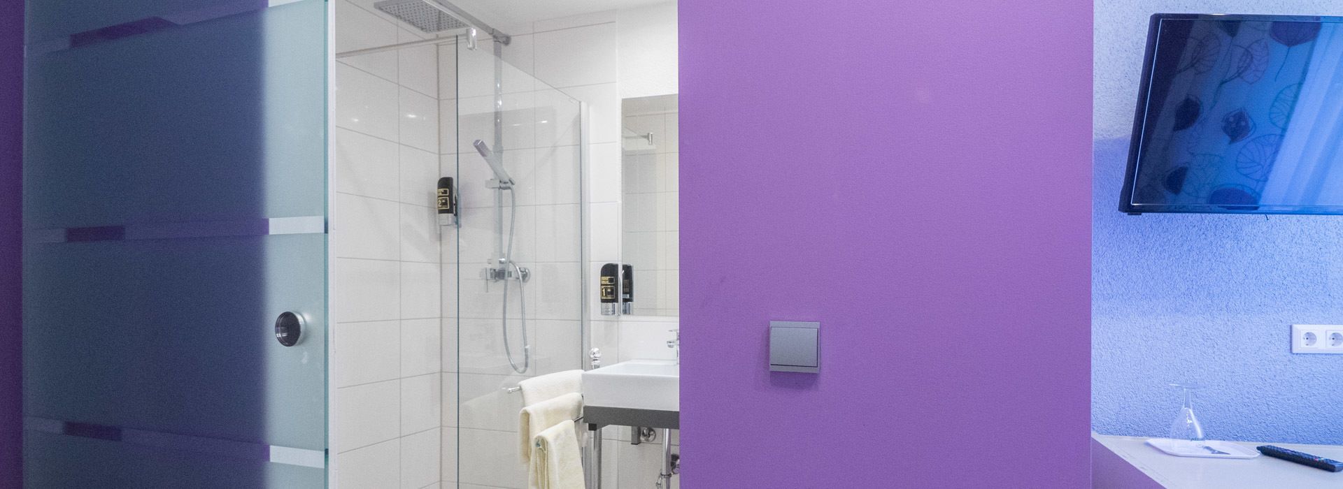 Un baño moderno con cabina de ducha de vidrio, un lavamanos blanco y una pared morada.