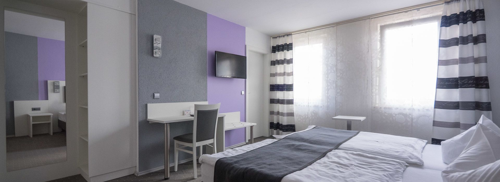 A hotel room with a bed, a desk, a chair, a TV, and a window with curtains.