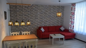 A living room with a red sofa, white chairs, a wooden dining table, hanging pendant lights, and a stone wall. - Hotel Holländischer Hof (Lich)