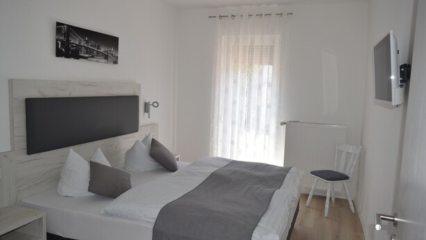 A modern bedroom with a large bed, a bedside table, a chair, a TV mounted on the wall, and a window with sheer curtains. - Hotel Holländischer Hof (Lich)