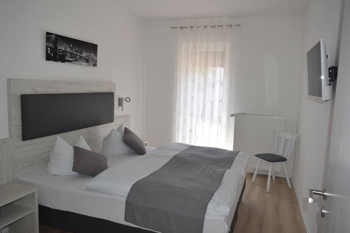 A modern bedroom with a large bed, a bedside table, a chair, a TV mounted on the wall, and a window with sheer curtains.