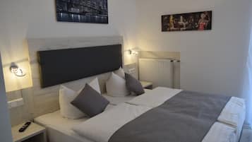 A neatly made bed with a grey blanket and white pillows, a nightstand with a remote control, and wall-mounted artwork depicting a cityscape and a painting of people.
