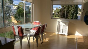 A dining area with a glass table and chairs, a white cabinet, and a view of greenery and buildings through large windows. - Spacious, comfortable, family home. Great views, pets ok, free wifi (Lakes Entrance)