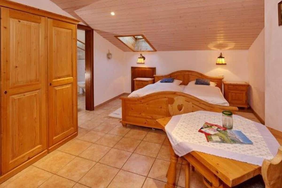 Ein Schlafzimmer mit einem hölzernen Bett, zwei Nachttischen, einem Schminktisch, einem Holzschrank, einem kleinen Tisch mit einer Decke und einem Oberlicht.