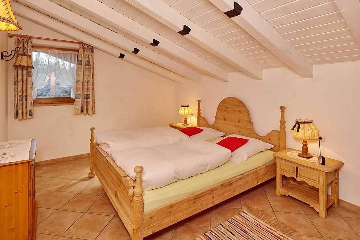 Ein Schlafzimmer mit einem hölzernen Bett, zwei Nachttischen mit Lampen, einem Fenster mit Vorhängen und einem kleinen Schreibtisch mit Lampe.