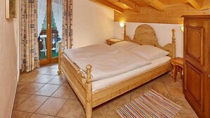 Una cama con estructura de madera, cabecera, mesita de noche y un armario en una habitación con suelo de baldosas y techo inclinado.