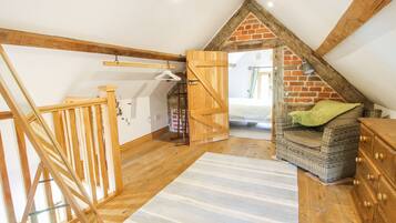 A loft bedroom with exposed wooden beams, a sloped ceiling, a bed, a wicker chair, and a wooden dresser.