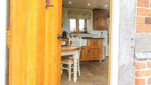 A wooden door leading into a kitchen with a dining table and chairs, wooden cabinets, and a brick wall. - The Shooting Folly (Market Drayton)