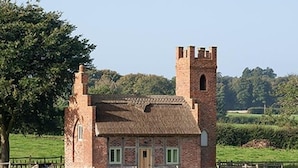 A rustic brick building with a tower, surrounded by a fenced pasture and a tree. - The Shooting Folly (Market Drayton)