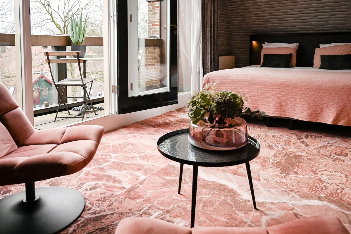 A modern bedroom with a bed, a chair, a small table with a plant, and a view of the outside.