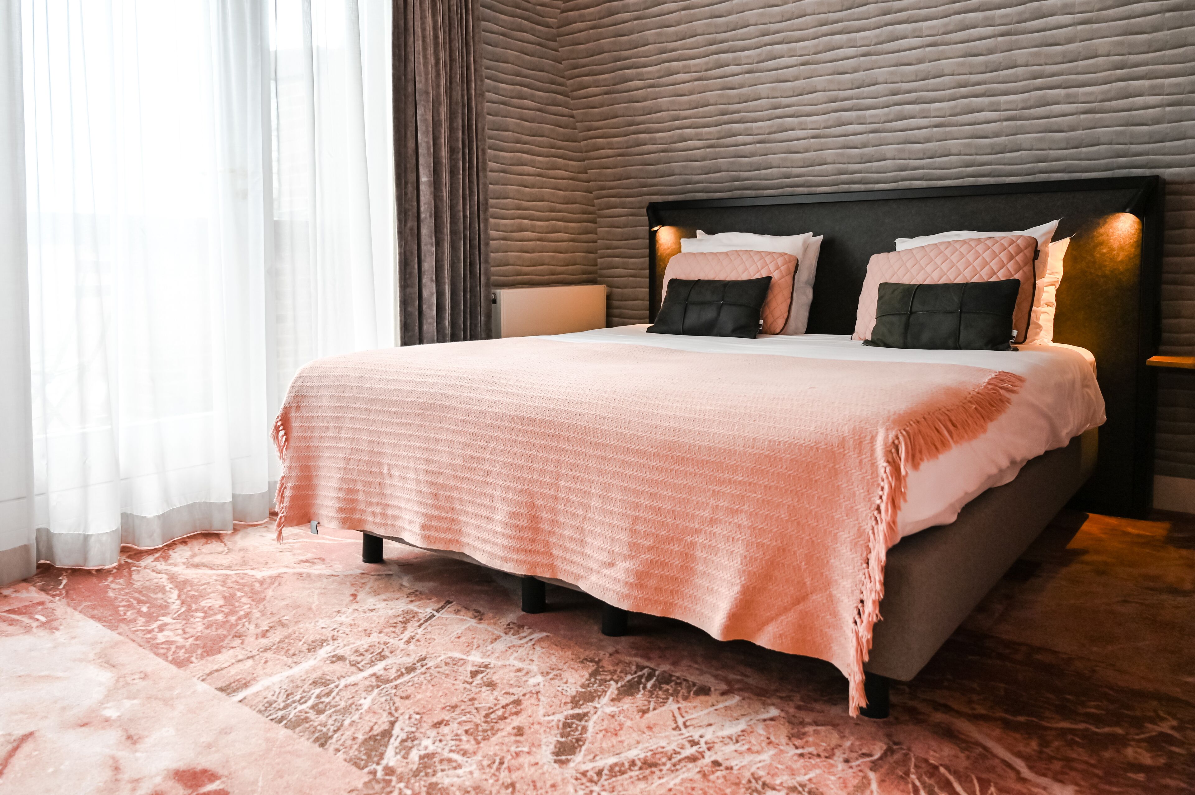 A neatly made bed with a pink bedspread and multiple pillows, a bedside table with a lamp, and a textured wall behind the bed.