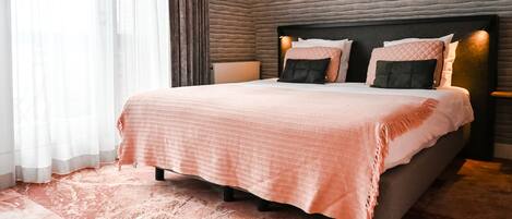 A neatly made bed with a pink bedspread and multiple pillows, a bedside table with a lamp, and a textured wall behind the bed.