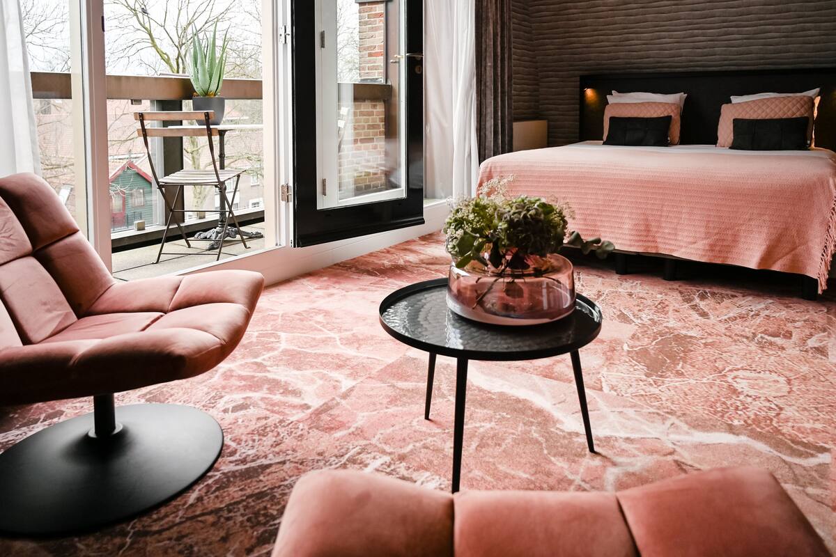 Ein Schlafzimmer mit einem Bett, einem Sessel, einem Tisch mit Pflanze und Blick durch ein Fenster.
