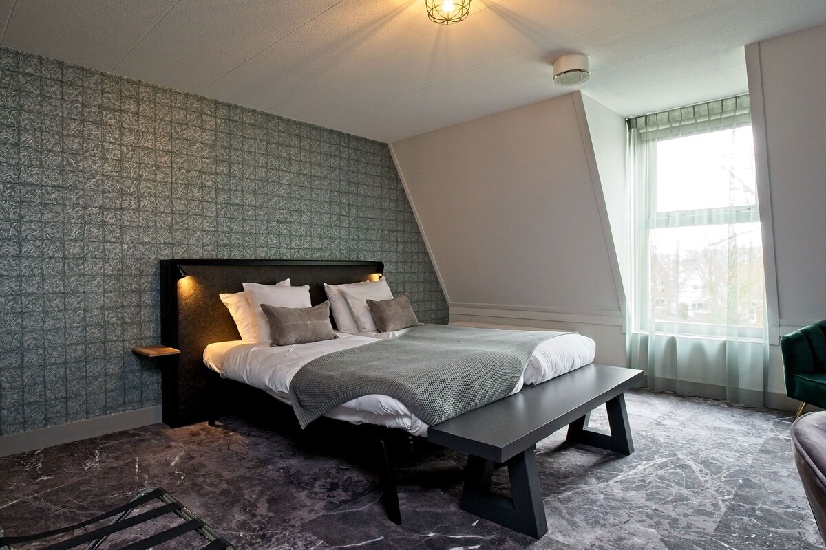 A modern bedroom with a bed, a bench, a nightstand, a lamp, and a window with curtains.