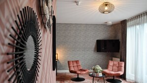 A modern living room with two pink armchairs, a round coffee table, a wall-mounted TV, and a unique wall decor. - Fletcher Slothotel Schagen (Schagen)