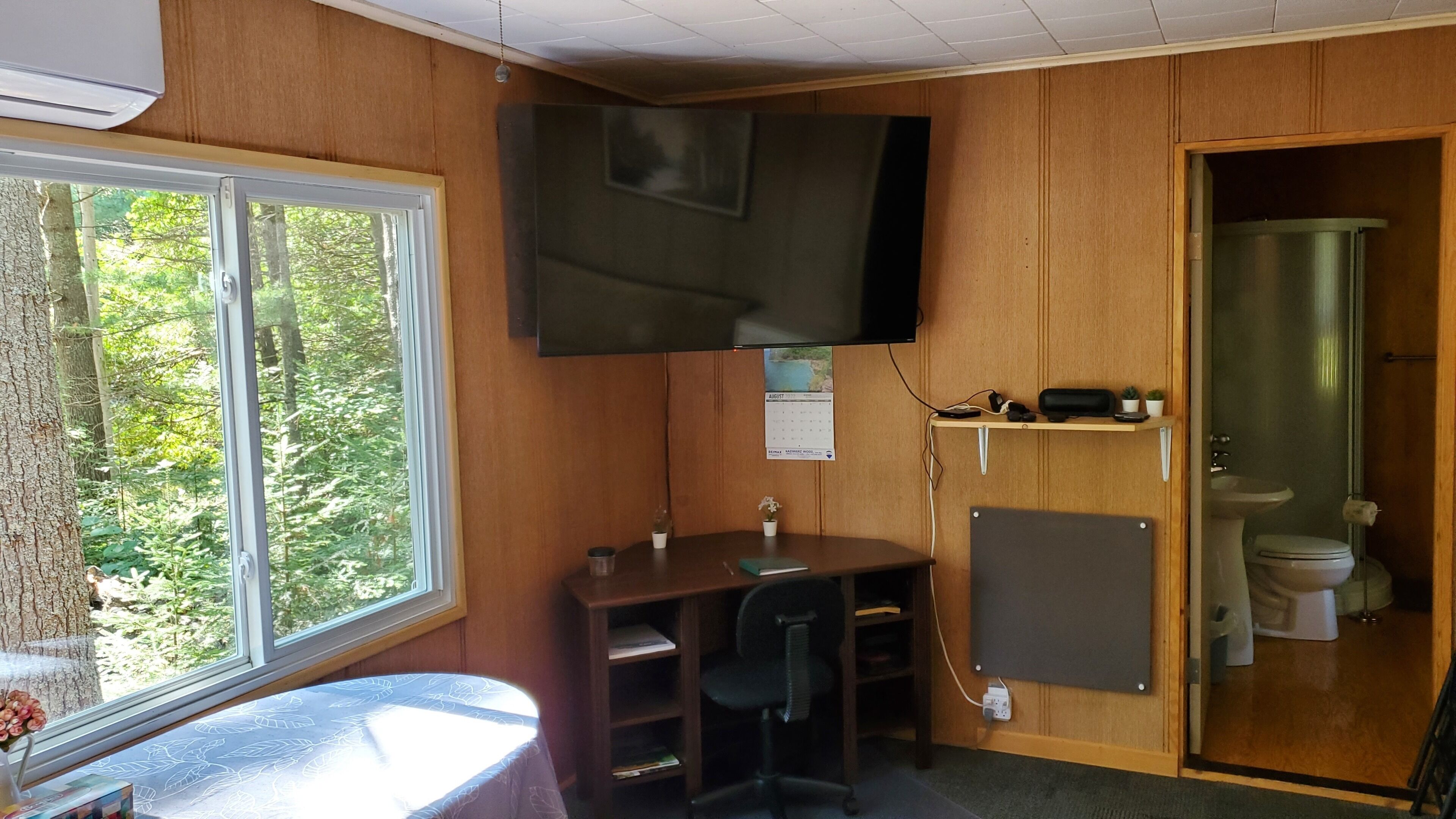 A compact room with a desk, chair, and a small shelf with personal items. A television is mounted on the wall, and a calendar is visible. A window offers a view of trees outside.