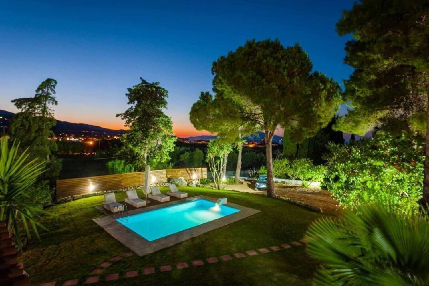 Zona de piscina exterior bien iluminada con sillones de descanso y vista al paisaje urbano al anochecer.