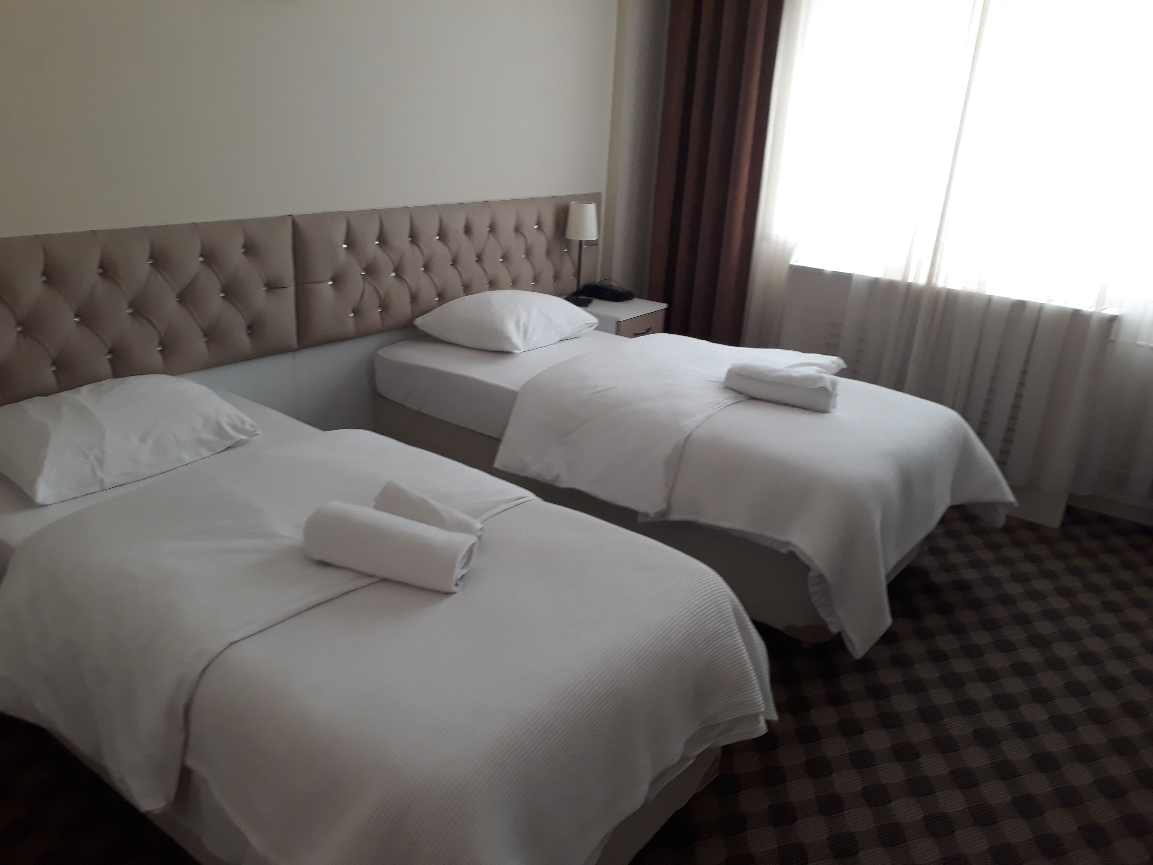 A hotel room with two beds, each with white linens and pillows, a tufted headboard, a nightstand with a lamp, and a window with curtains.