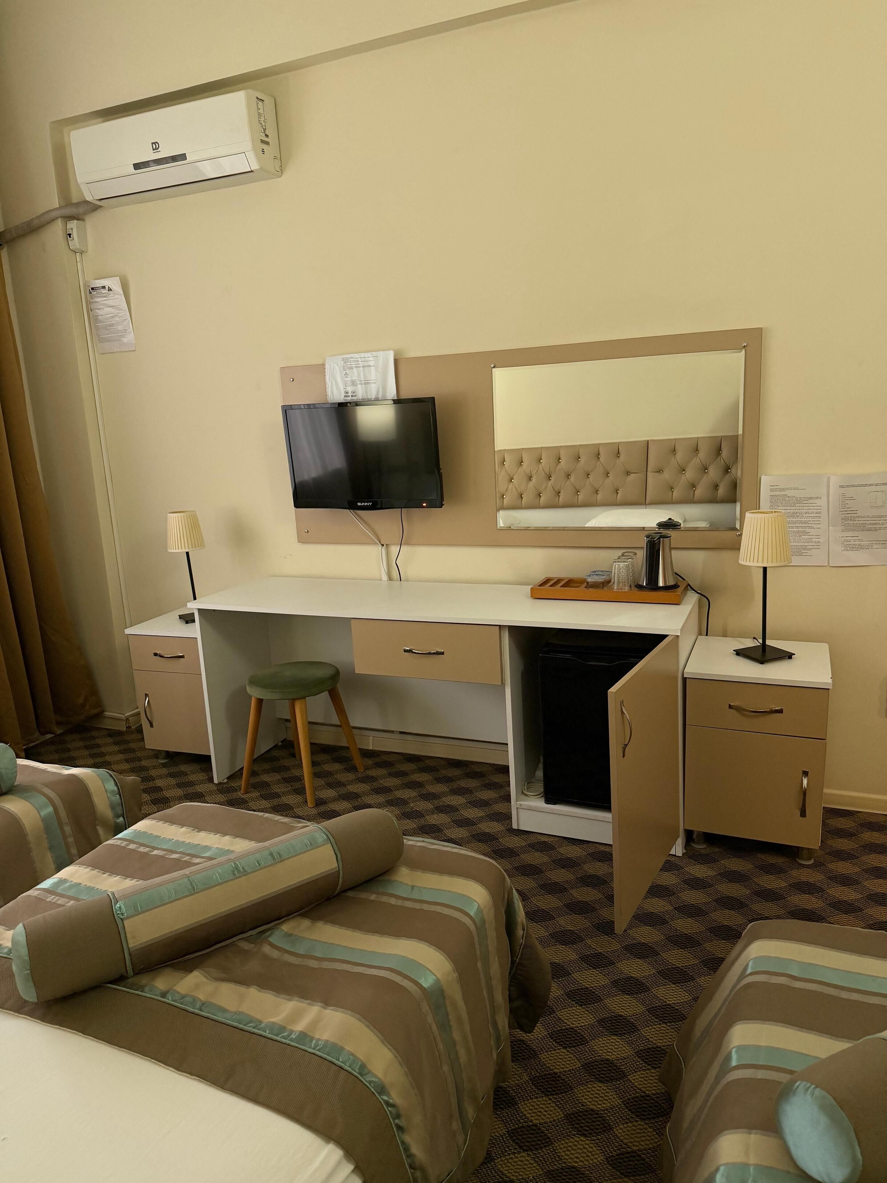 A hotel room with a bed, a desk with a television, a minibar, and a large mirror.
