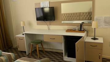 A hotel room with a bed, a desk with a television, a minibar, and a large mirror.