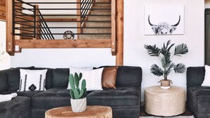 Ein modernes Wohnzimmer mit einem dunkelgrauen Ecksofa, einem Holztisch und einer Kakteenpflanze. Im Hintergrund ist eine Treppe mit Metallgeländer zu sehen.