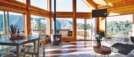 A cozy cabin interior with a dining area, a sofa, and a fireplace. Large windows offer a view of snow-covered mountains.