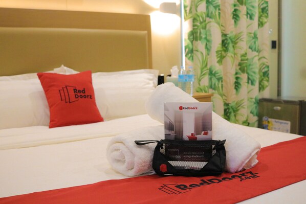 A neatly made bed with a red pillow, a rolled white towel, and a RedDoorz branded bag.