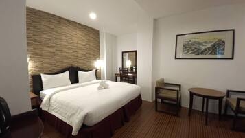 A hotel room with a large bed, a wooden headboard, a desk, a chair, a small table, and a framed artwork on the wall.