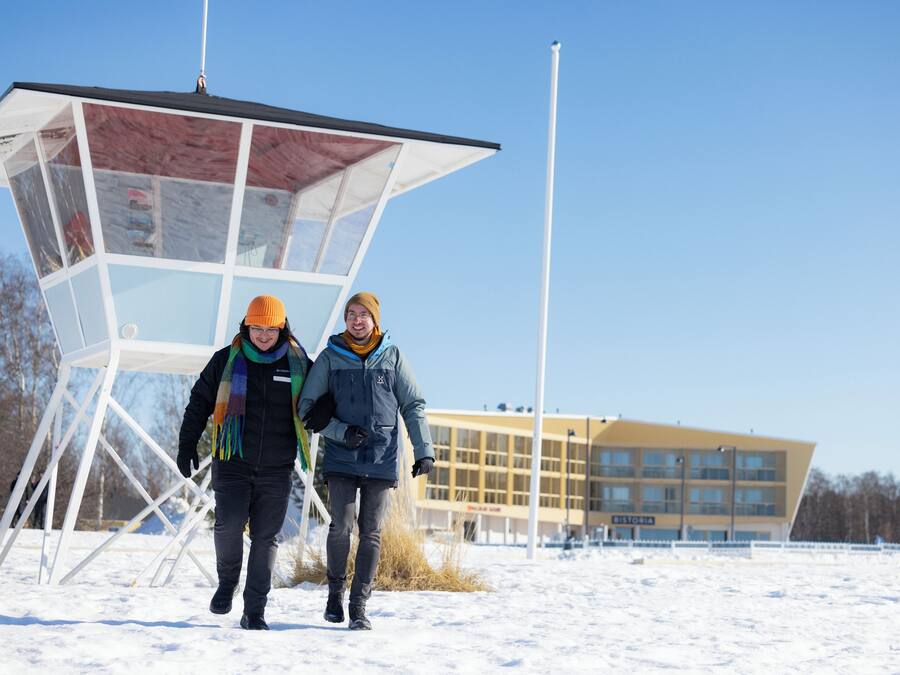 Two people walking in a snowy area with a modern building and a glass structure in the background.