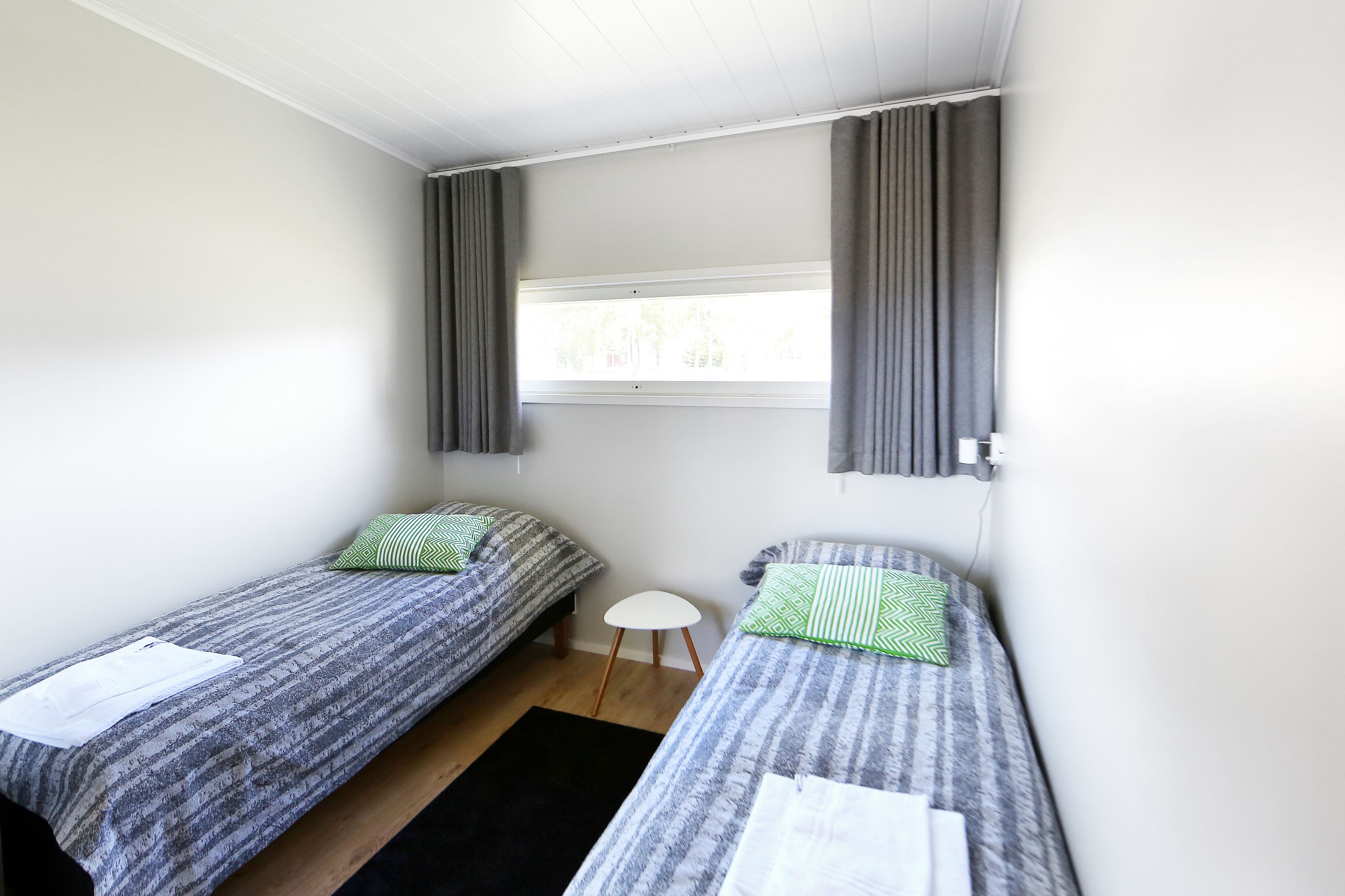A small, clean room with two single beds, a small white table, and a window with curtains.