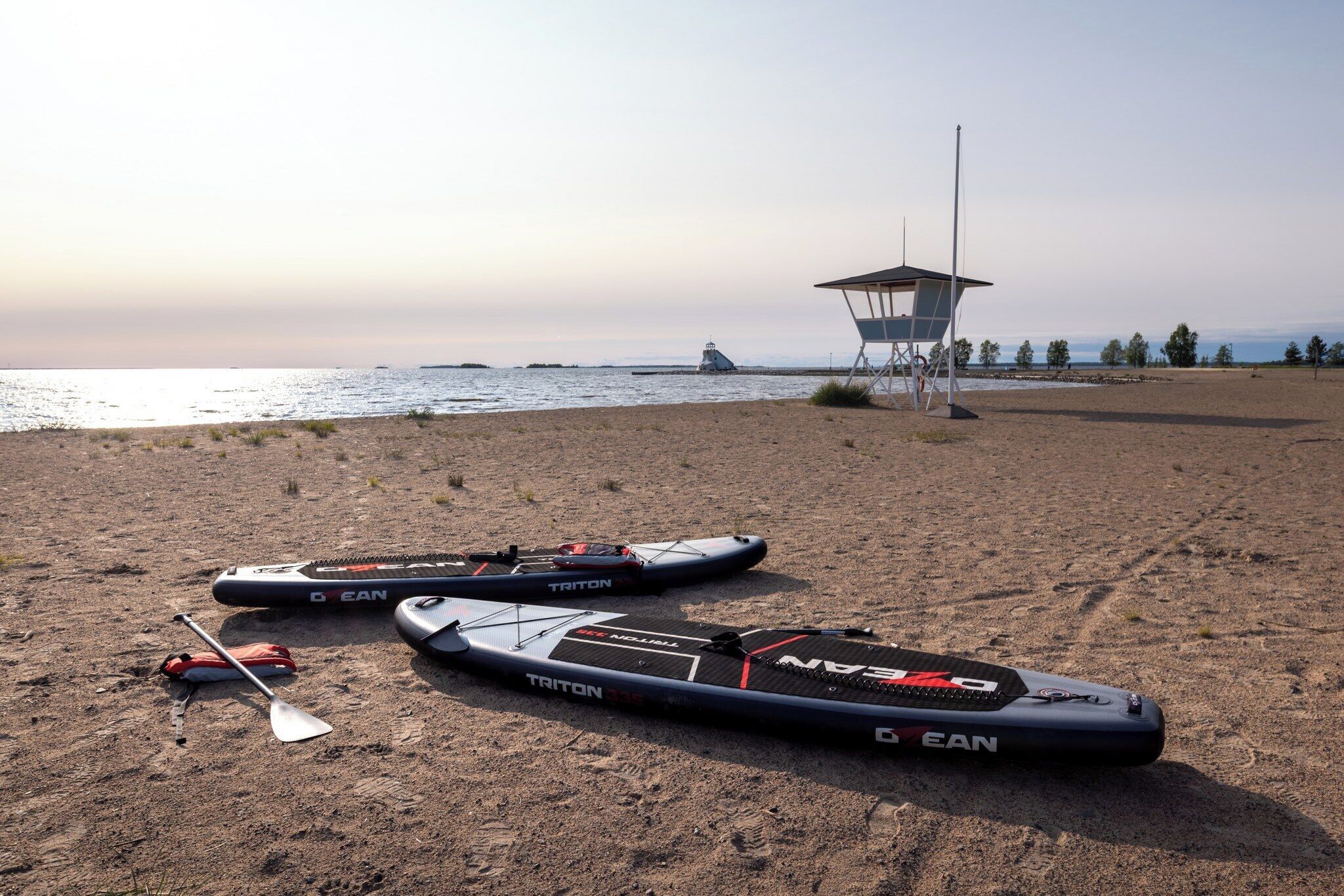 A beach with two Triton paddleboards, a paddle, and a lifeguard tower.