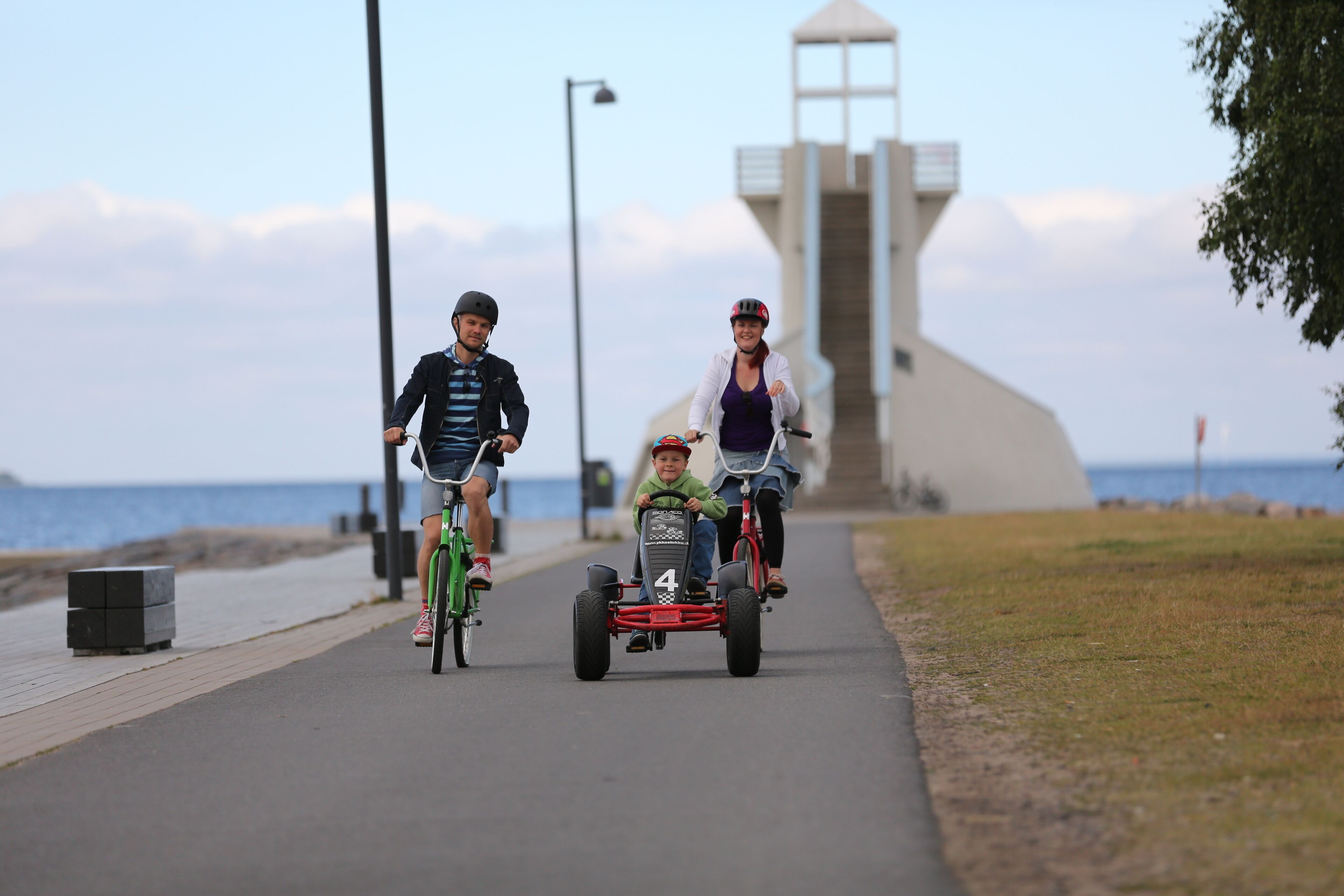 Tres personas paseando en karts sobre un camino pavimentado, con un faro al fondo.