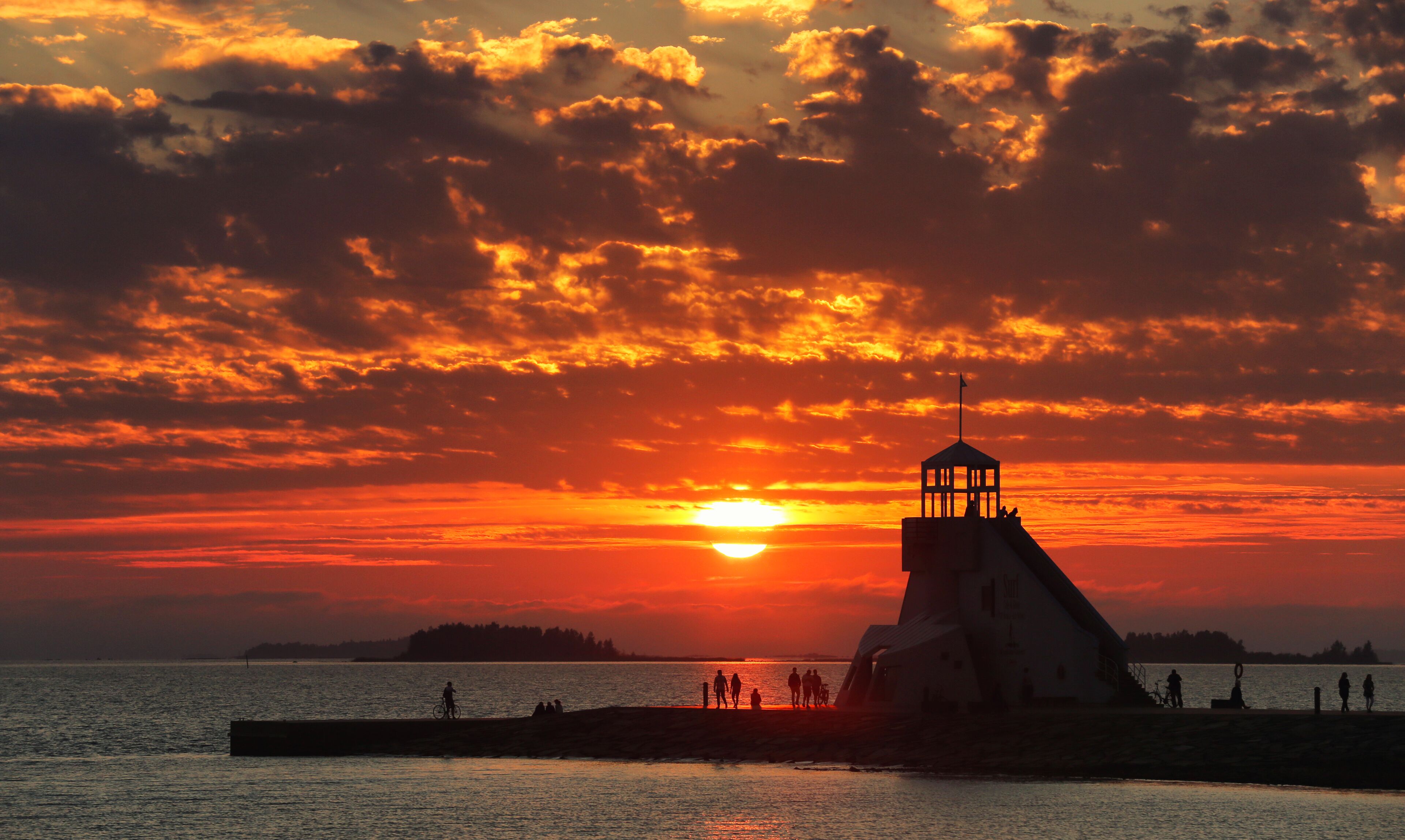 A lighthouse silhouette against a sunset with people and a bicycle on the pier.