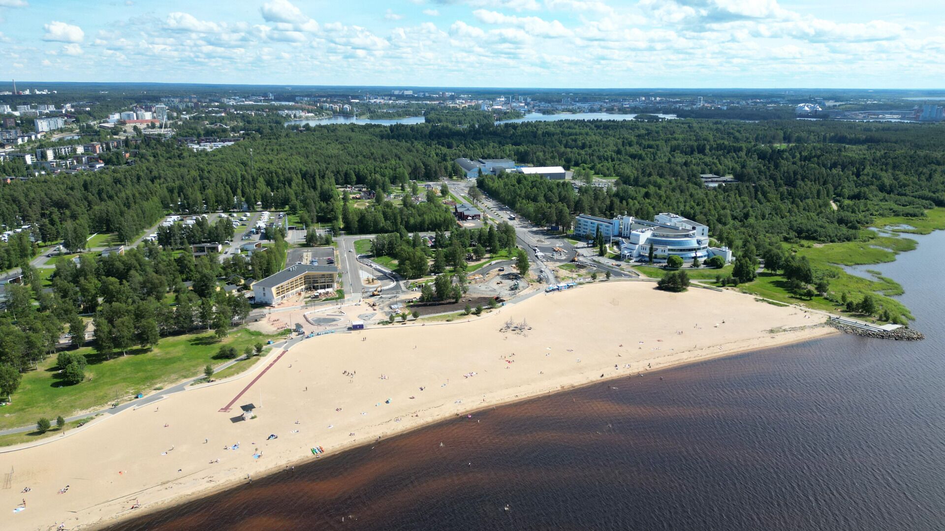 An aerial view of a coastal area with a sandy beach, a marina, and a mix of residential and commercial buildings.