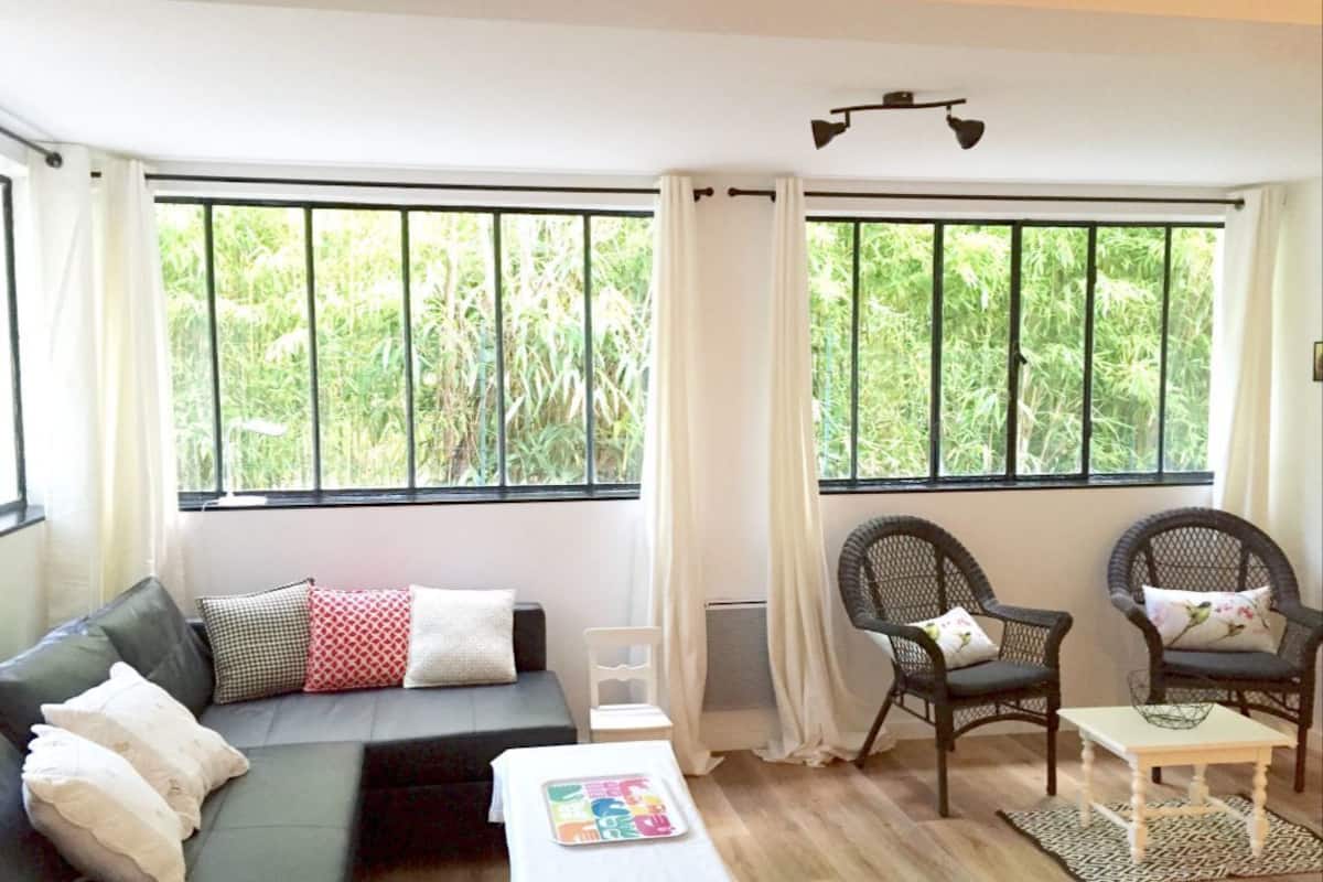 A cozy living room with a sofa, two wicker chairs, a small table, and large windows with curtains.