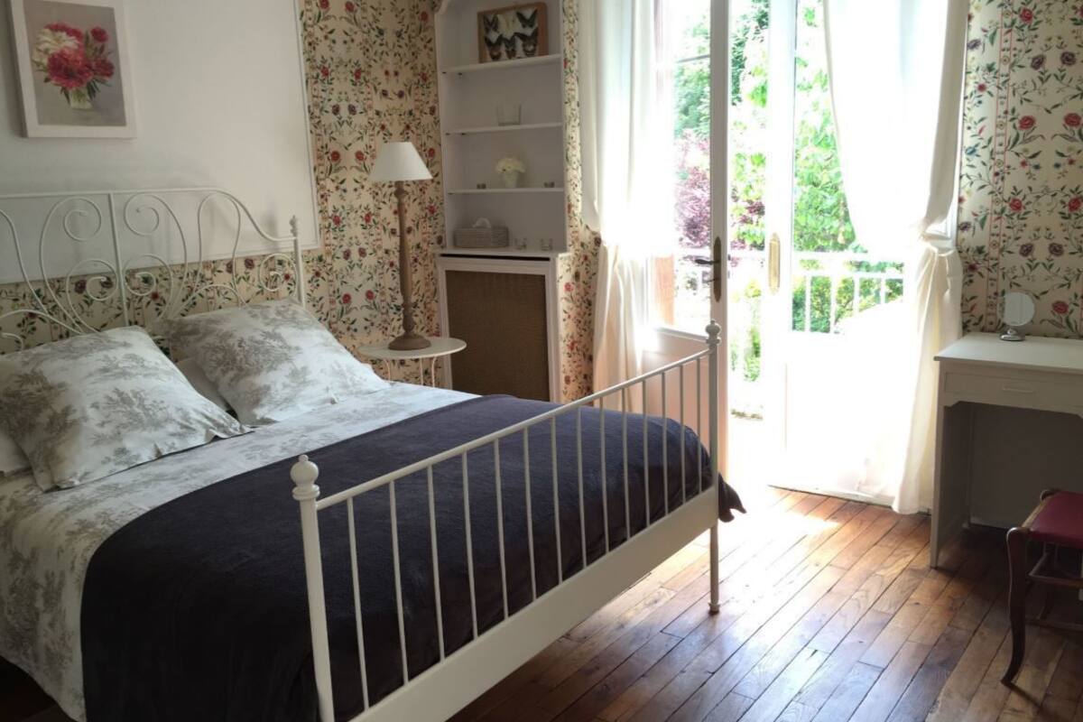 A bedroom with a bed, a nightstand, a lamp, a shelf, and a window with a view of greenery.