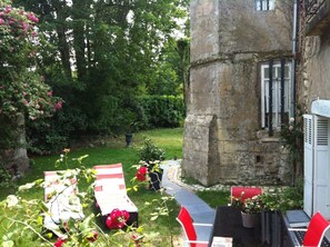A stone building with a narrow entrance, surrounded by lush greenery and a pathway leading to a seating area with red chairs and a table.