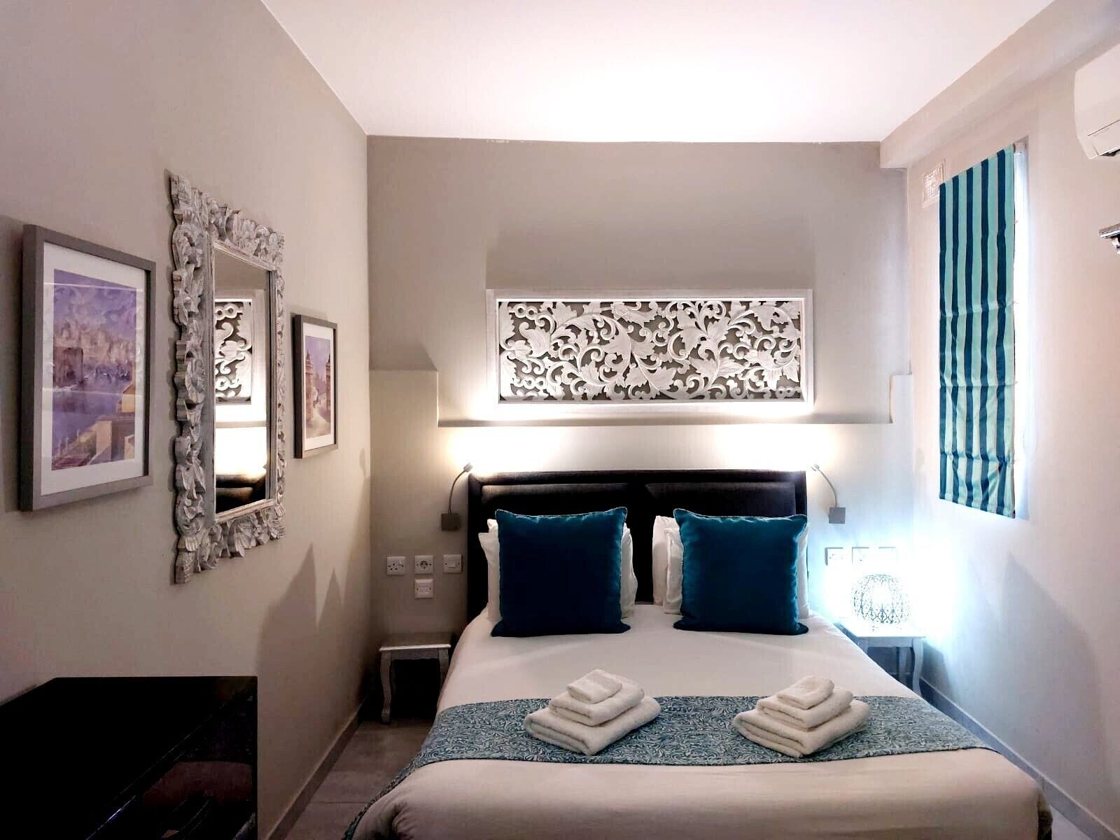 A hotel room with a large bed, two pillows, a decorative wall panel, a mirror, and a window with curtains.