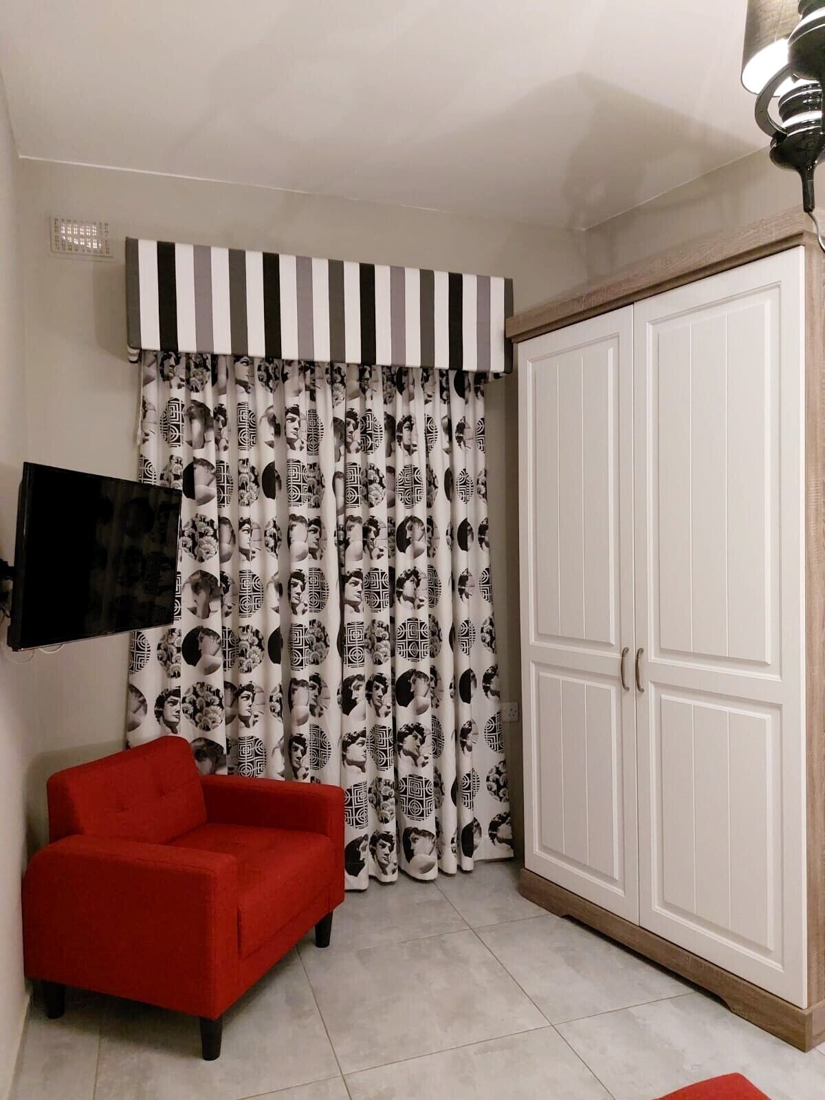 Una habitación con un sillón rojo, un armario blanco y una cortina con un estampado de rostros.