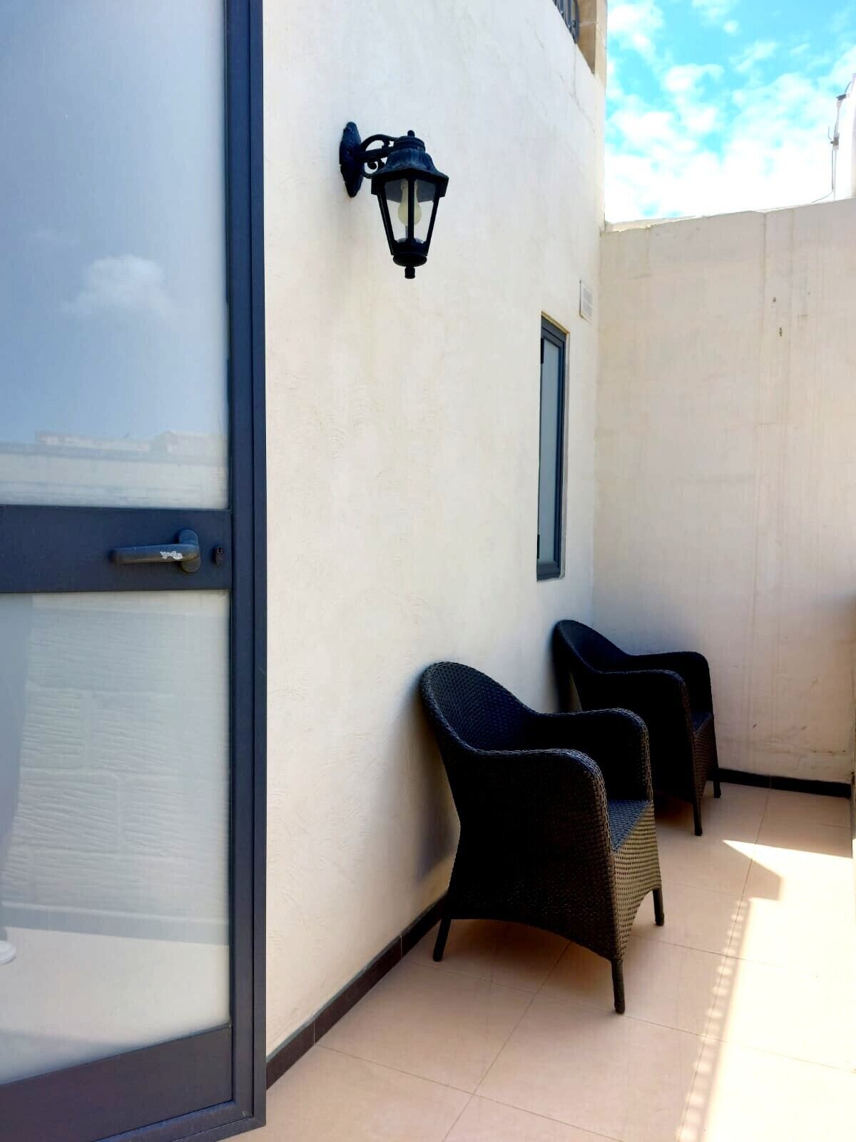 Un balcón con dos sillones de mimbre, un farol en la pared y una puerta con panel de vidrio.