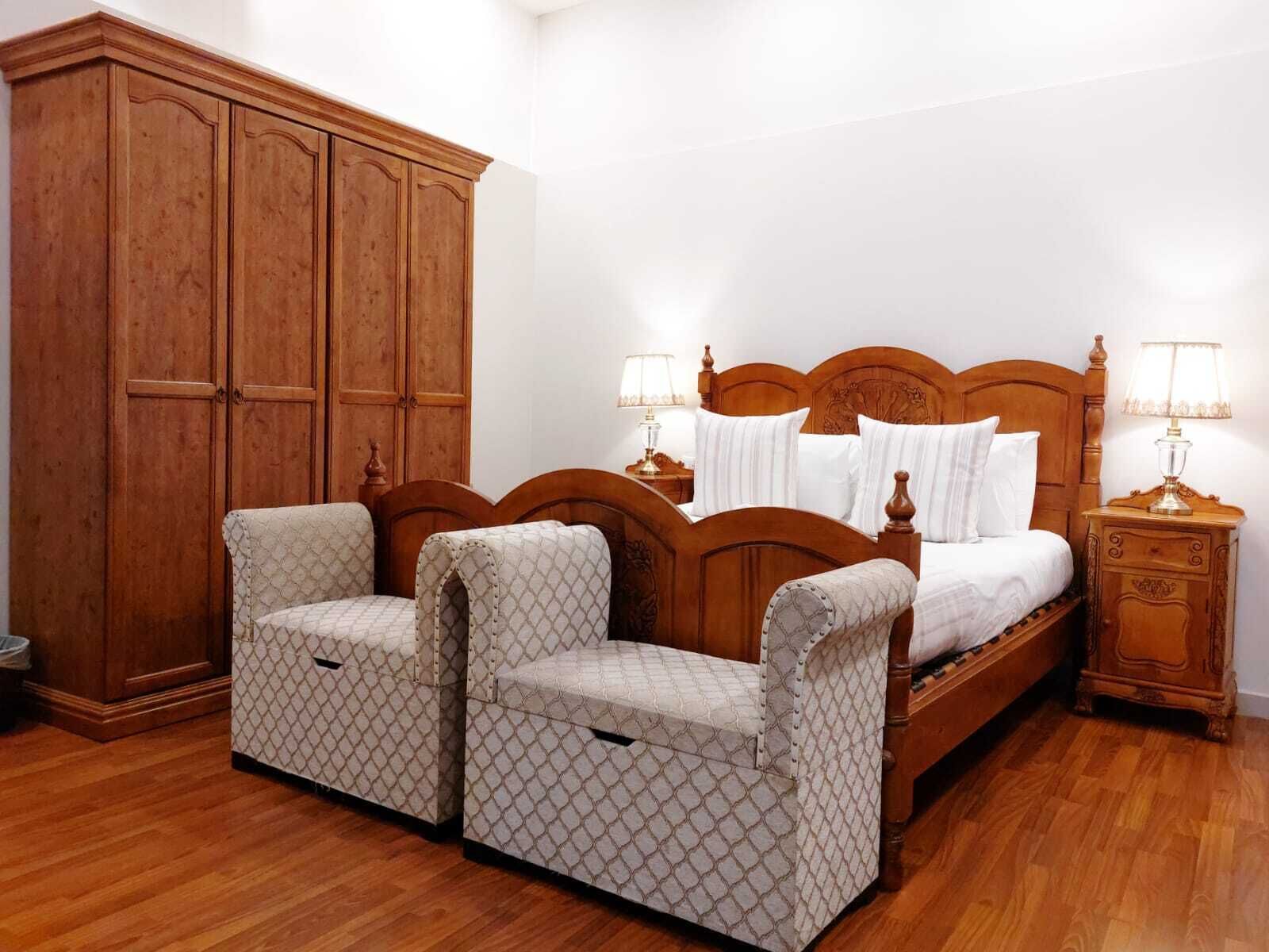 A bedroom with a wooden bed, two bedside tables with lamps, a large wooden wardrobe, and two patterned armchairs.