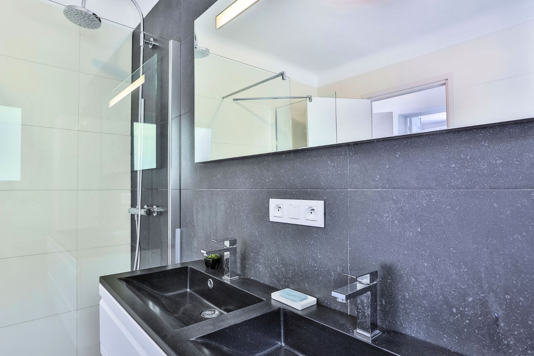 A modern bathroom with a glass shower enclosure, a black sink with a soap dish, and a mirror above the sink.