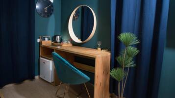 A compact room with a wooden desk, a teal chair, a small white fridge, a mirror, a clock, and a potted plant.
