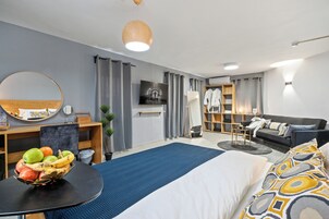 Una habitación de hotel moderna con cama, espejo redondo, una mesa redonda con fruta, televisor, armario y una zona de estar con sofá y una mesita.