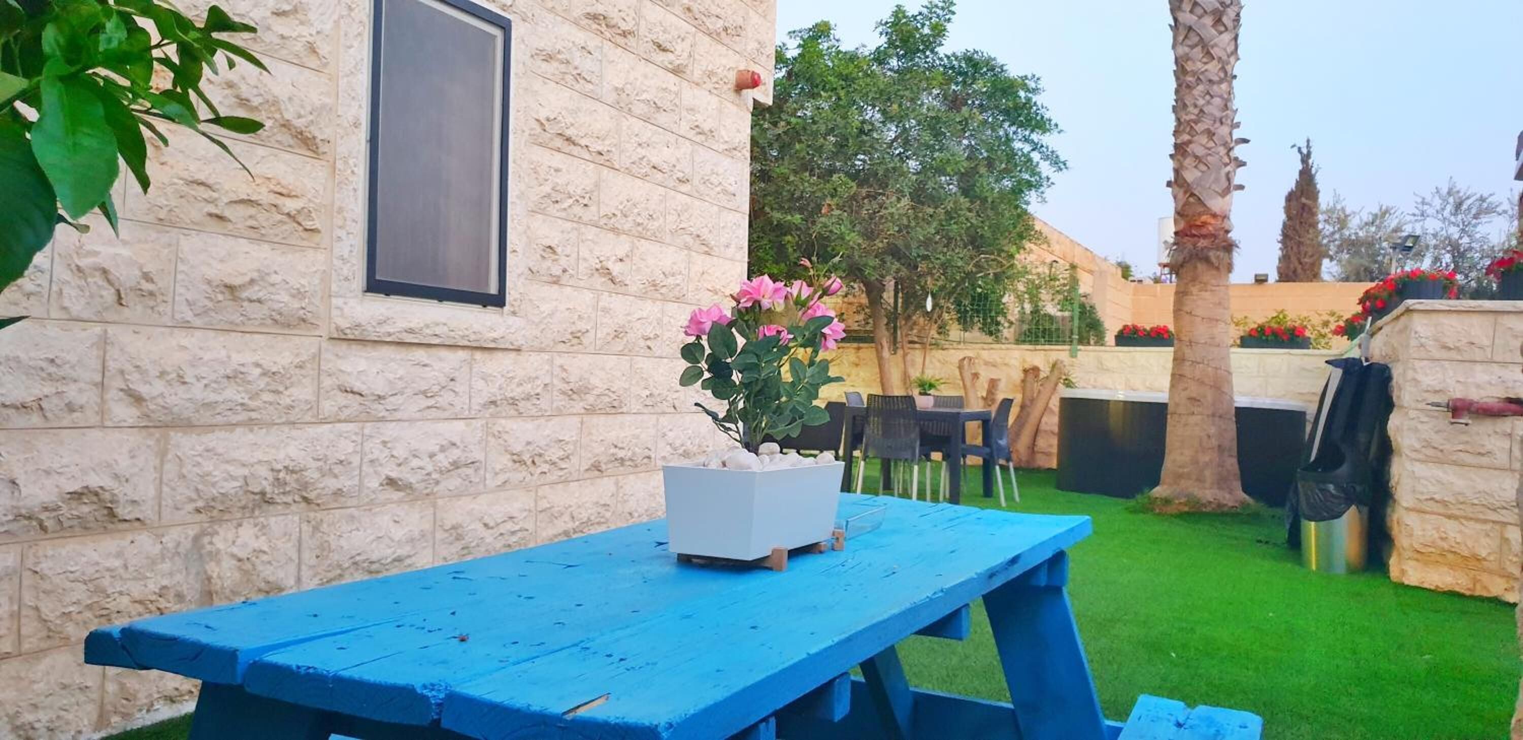 A stone building with a window, a blue table with a flower pot, and a green lawn with trees and chairs.