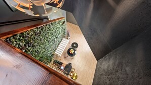 Een ruimte binnenshuis met een houten vloer, een groene wand vol planten, een zithoek met een tafel en stoelen, en een moderne lamp.