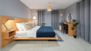 Een moderne slaapkamer met een houten hoofdbord, een bed met een blauwe spreideken, een bureau met een stoel, een ronde spiegel en een kleine tafel met een lamp.