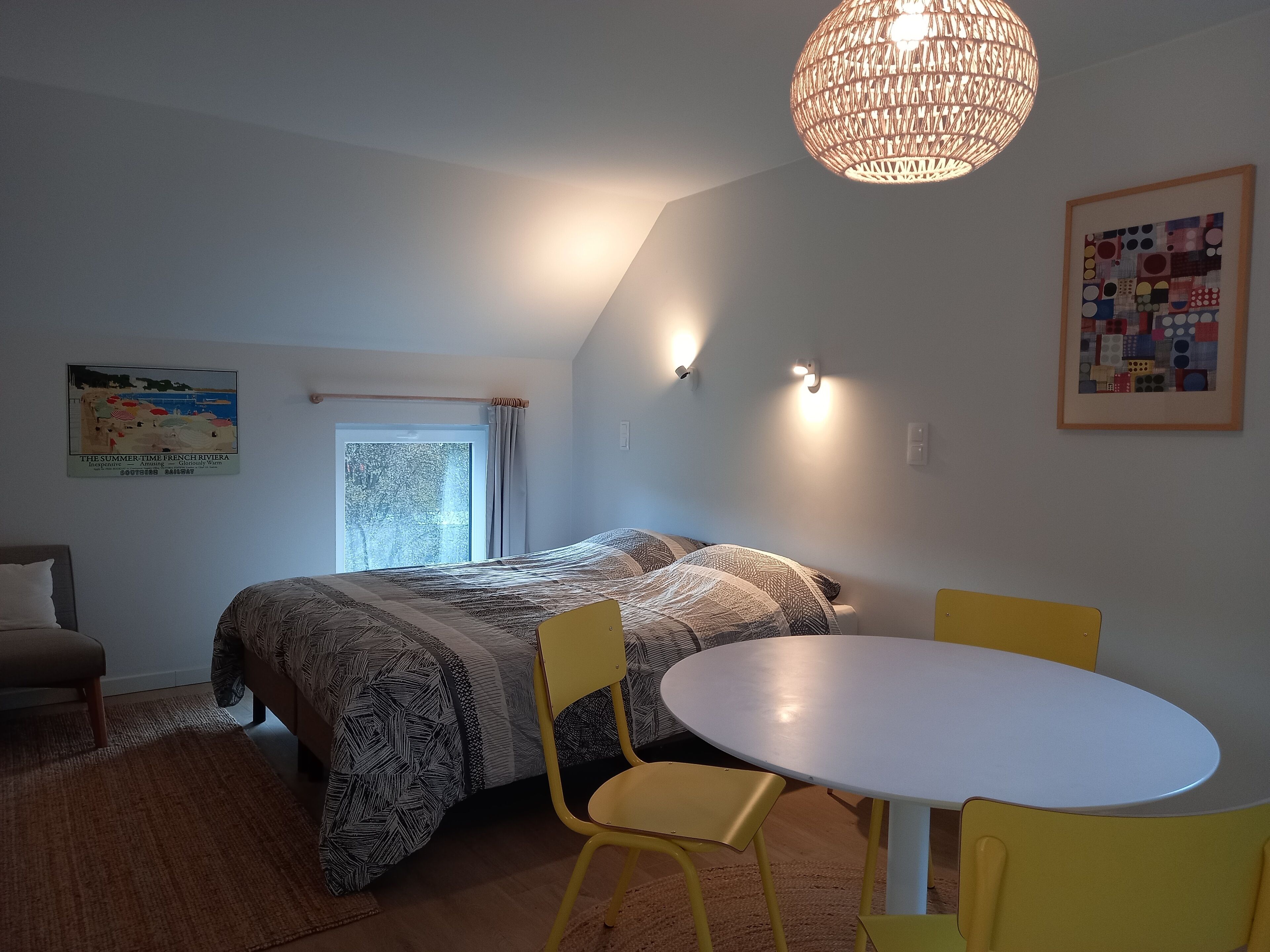 A bedroom with a bed, a round table, and two chairs. There is a window with curtains, a wall-mounted light, and a framed picture.