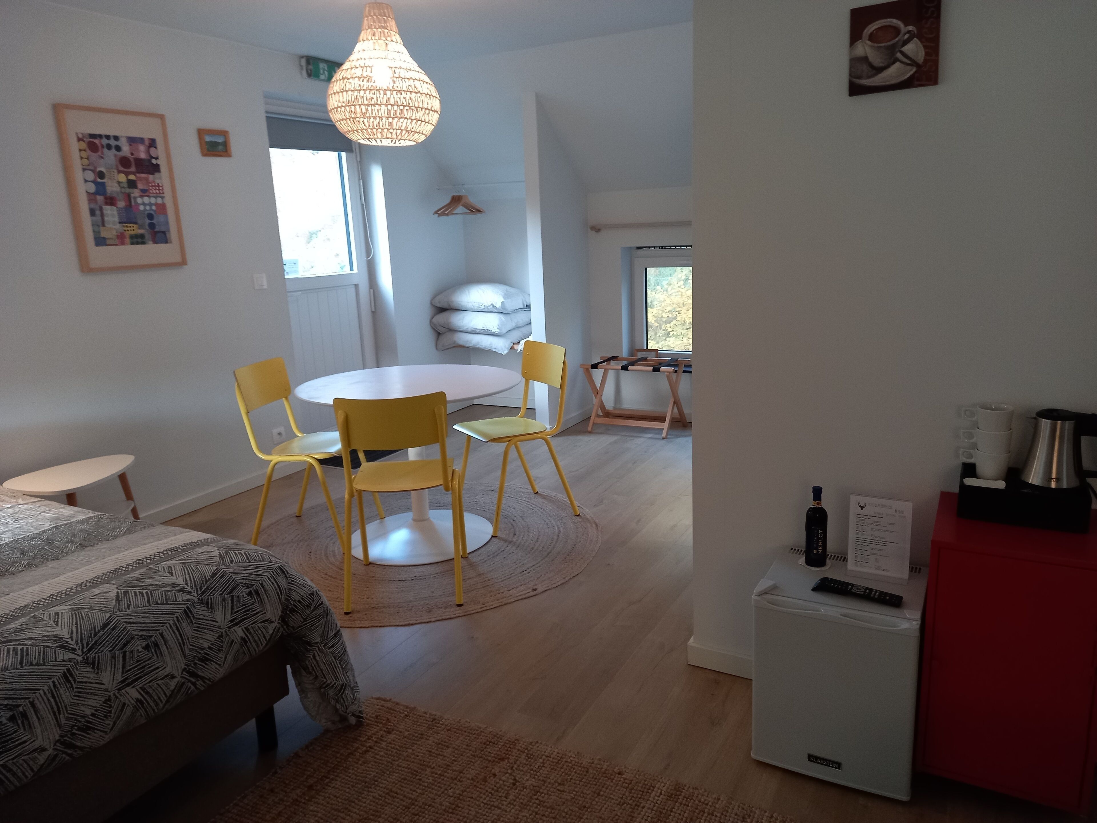 Een moderne, goed verlichte kamer met een ronde eettafel, gele stoelen, een bed met een beddengoed met dessin, een kleine witte koelkast en een koffieapparaat.